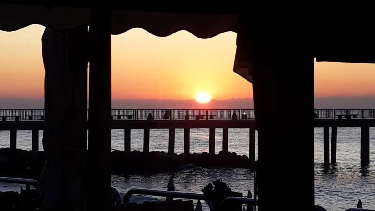 Sole basso sul mare e pontile, visti dalla terrazza coperta dello stabilimento balneare.