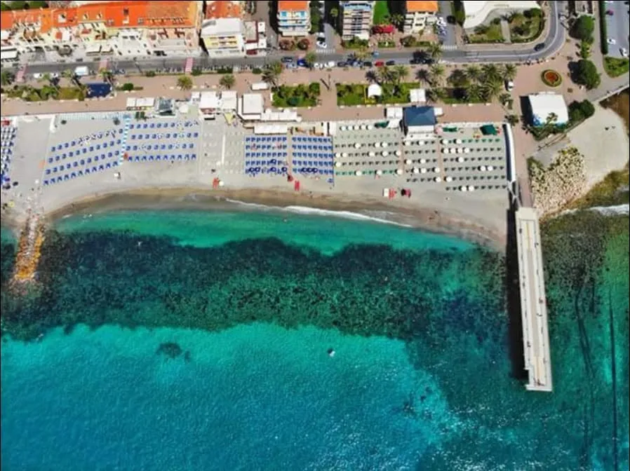 Vista aerea della spiaggia con ombrelloni allineati, mare turchese, pontile e passeggiata lungomare.