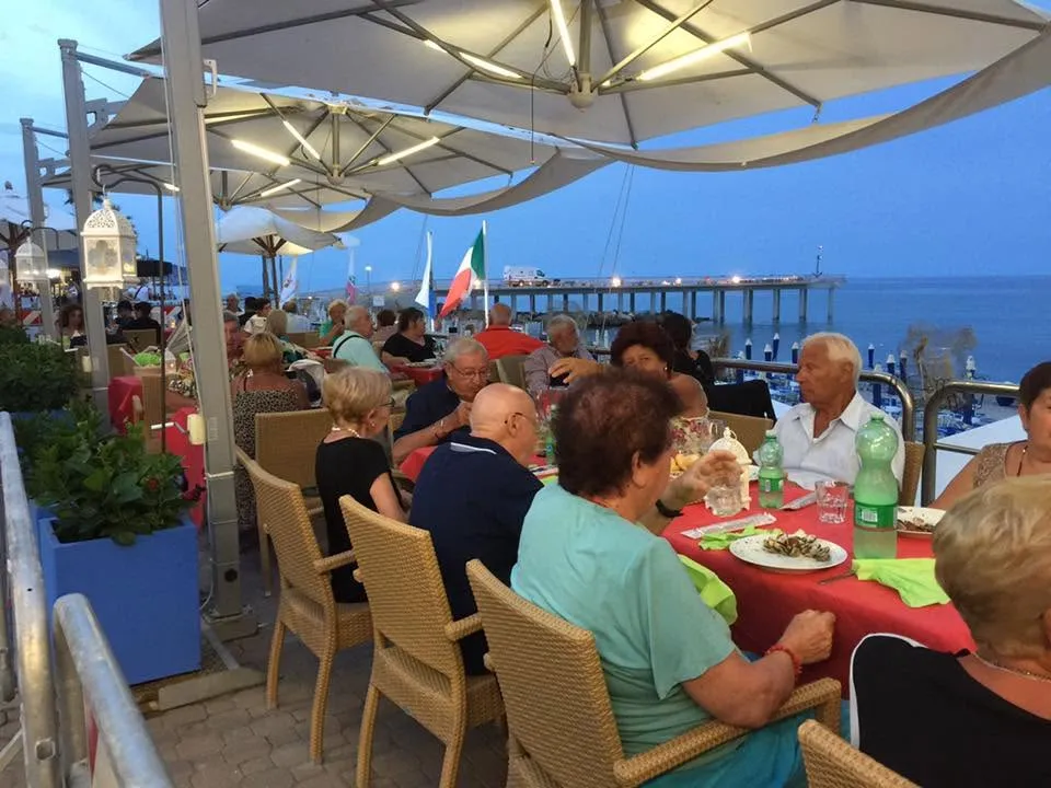 Persone a cena sulla terrazza sul mare, sotto ombrelloni illuminati, con pontile sullo sfondo.